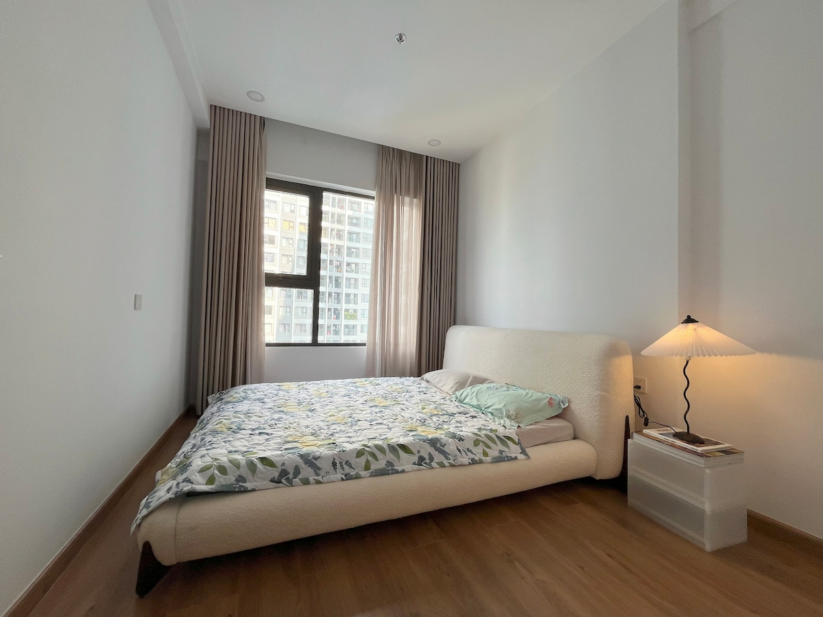 A spacious sleeping area features a low-profile bed adorned with a floral-patterned blanket and soft pillows. Natural light enters through large windows, surrounded by sheer curtains. A bedside table holds a modern lamp, complementing the neutral color palette of the room.