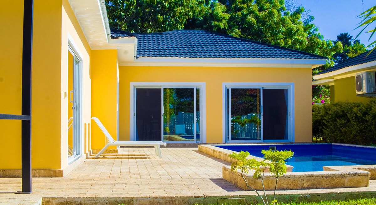 The villa exterior features a vibrant yellow facade, complemented by large glass windows reflecting lush greenery. A sleek lounge chair is positioned beside a rectangular pool, which is bordered by a stone patio. The surrounding landscape includes a well-maintained lawn and tropical plants.