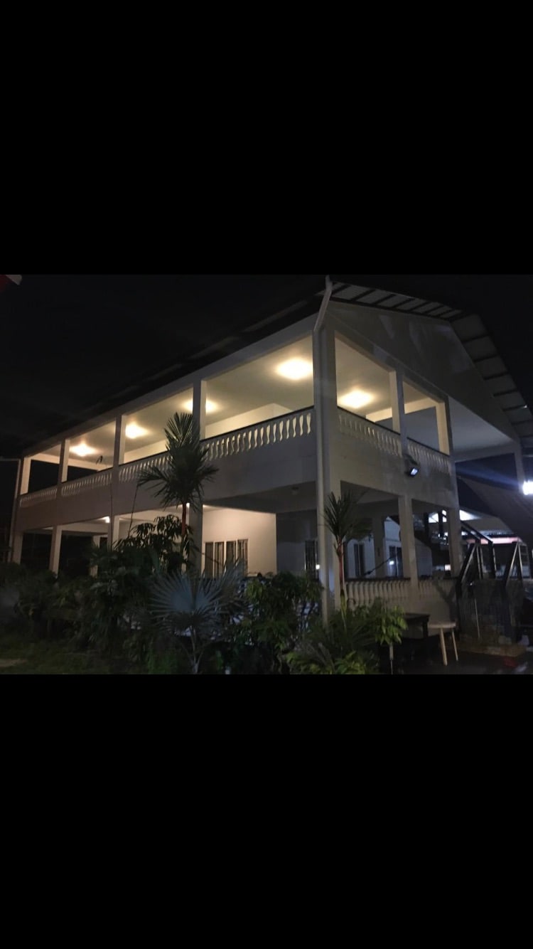 A two-story building is illuminated by soft lighting, highlighting its architectural features. A spacious balcony runs along the front, adorned with a railing and surrounded by lush greenery. The outdoor area includes various plants, creating a calm atmosphere.