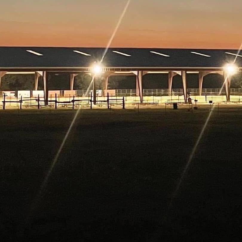 A covered arena is illuminated at dusk, showing its spacious interior designed for riding and events. Bright lights reflect off the structure, while the surrounding landscape is visible in the soft evening glow.
