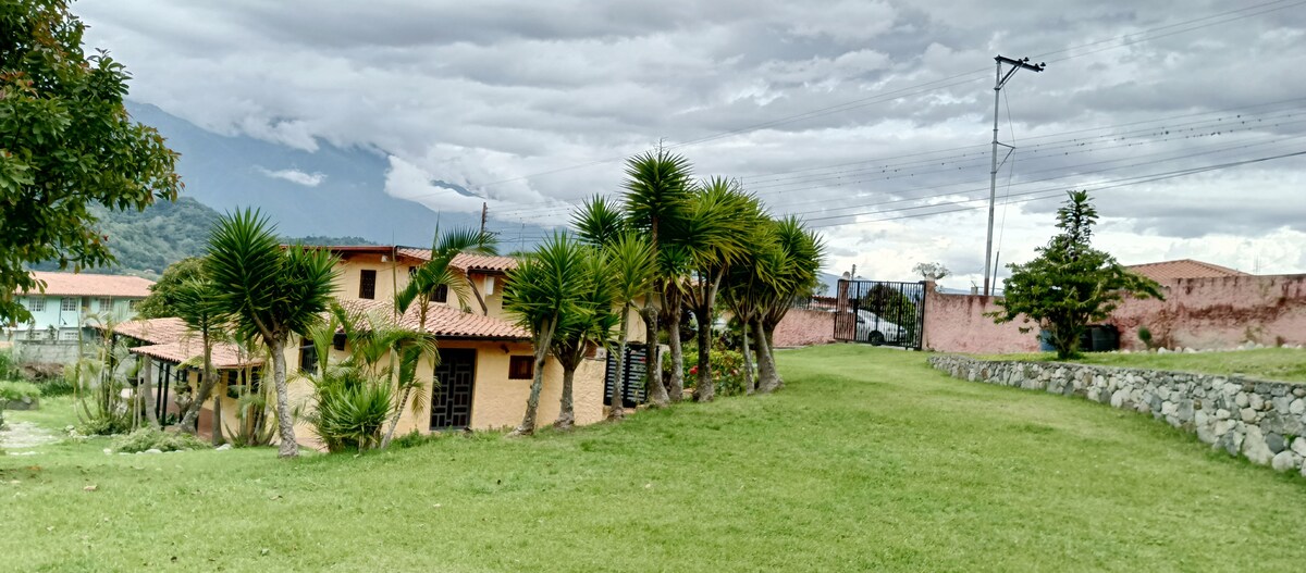 A lush green lawn surrounds a single-story building with a terracotta roof, flanked by palm trees. A scenic mountain backdrop can be seen in the distance, under a cloudy sky. The area appears spacious and well-maintained, with a stone wall subtly outlining the property.