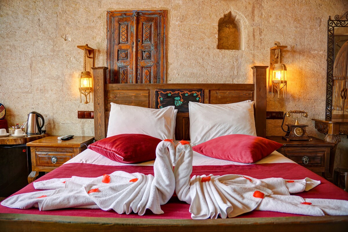 A spacious bed adorned with soft, white linens, accented by vibrant red pillows. Decorative towels are elegantly arranged on the bed, contributing to a cozy atmosphere. The room features unique stone walls, along with wooden accents and antique-style lighting for a charming ambiance.