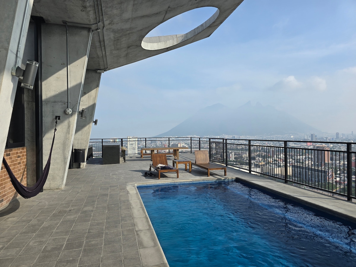 An outdoor terrace features a pool surrounded by a sleek tiled floor. Two wooden lounge chairs are positioned nearby, offering scenic views of the distant mountains and city skyline. A hammock is mounted from the structure's wall, inviting relaxation in the open air.