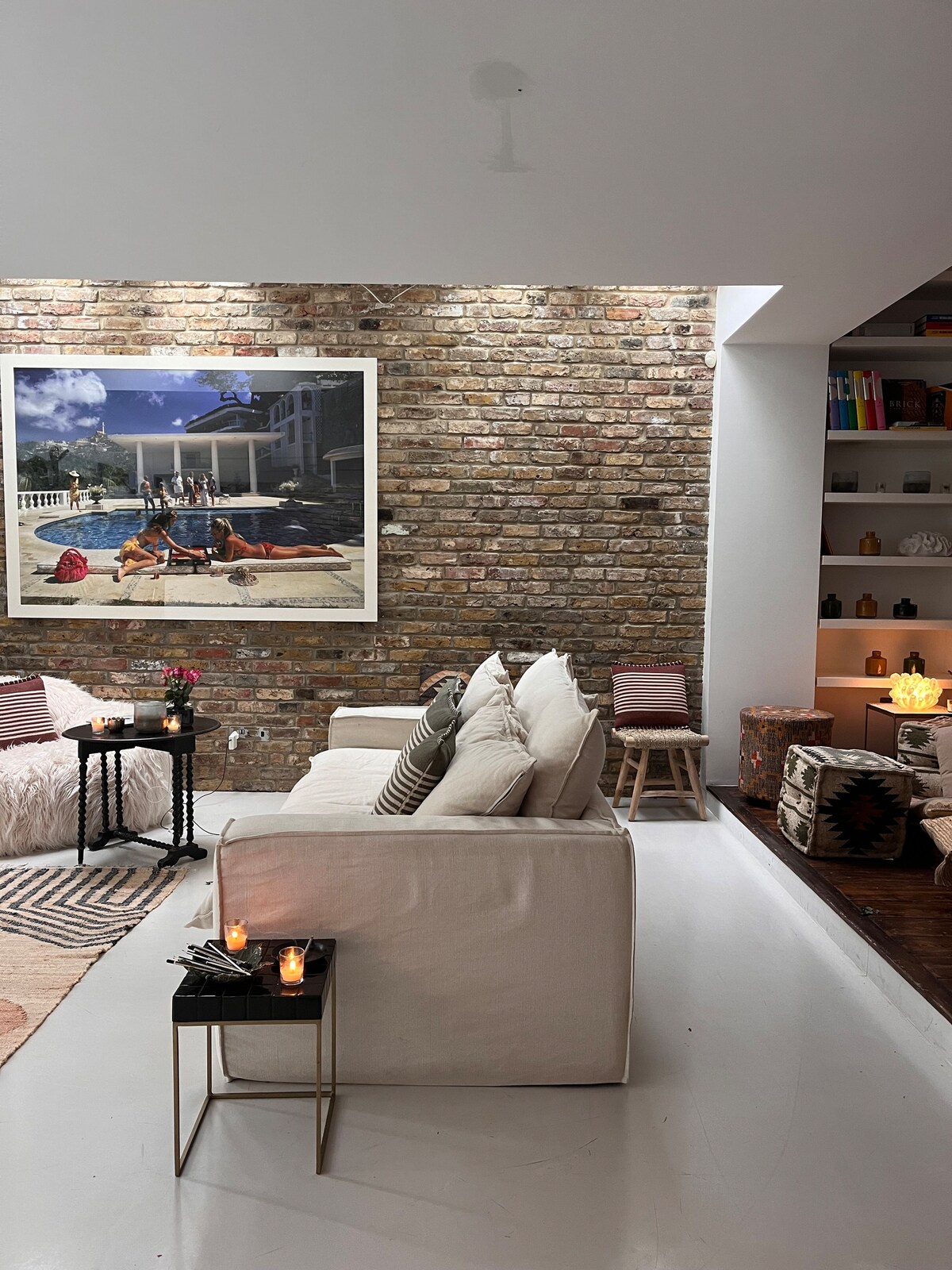 The living area features a white, spacious sofa set against a rustic brick wall, creating a cozy ambiance. A large art piece hangs above, depicting a lively outdoor scene. Small side tables with candles provide a warm glow, enhancing the room's inviting atmosphere.