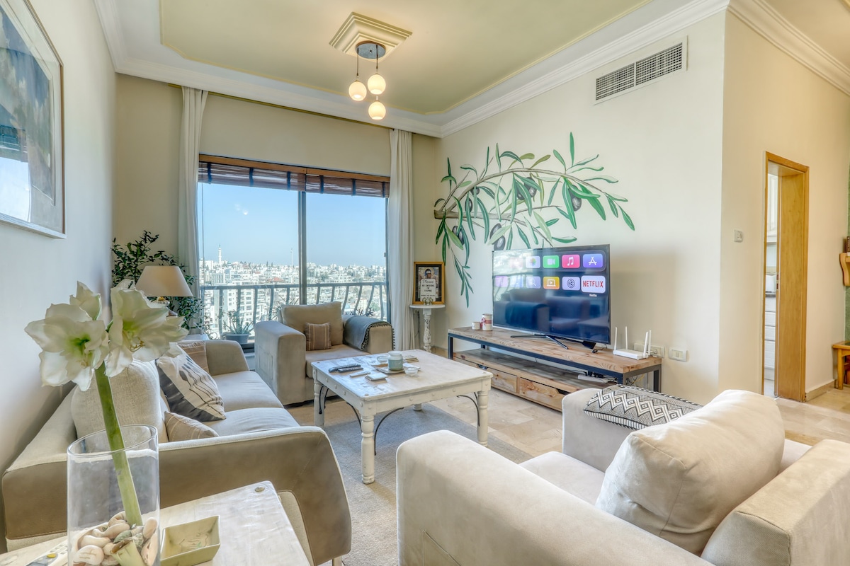 A cozy living room is featured, showcasing two beige sofas facing a coffee table. A television is mounted on the wall, displaying a variety of streaming options. Large windows offer a view of the city skyline, and a mural of greenery adds a touch of nature to the space.