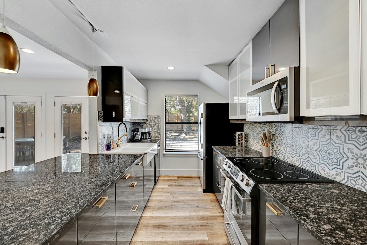 A modern kitchen features sleek cabinetry in black and white, complemented by granite countertops. Stainless steel appliances are prominently displayed, including an oven and microwave. Natural light streams through large windows, highlighting the stylish backsplash design.