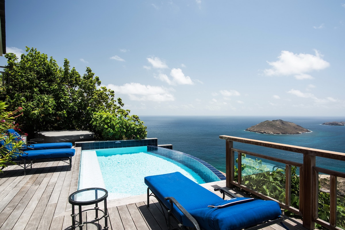 An outdoor area features a private pool surrounded by wooden decking. Two lounge chairs in a deep blue hue are positioned nearby, overlooking a stunning ocean view that includes distant islands and gentle waves. Lush greenery frames the scene, enhancing the serene atmosphere.