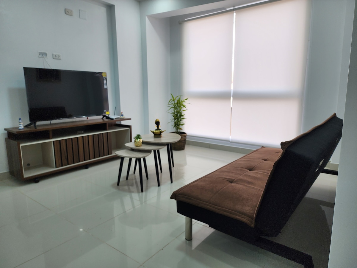 A modern living area features a comfortable sofa bed positioned across from a flat-screen TV mounted on a wooden entertainment unit. Two small coffee tables are placed in front, with a potted plant adding a touch of greenery beside a large window covered by light-filtering blinds.