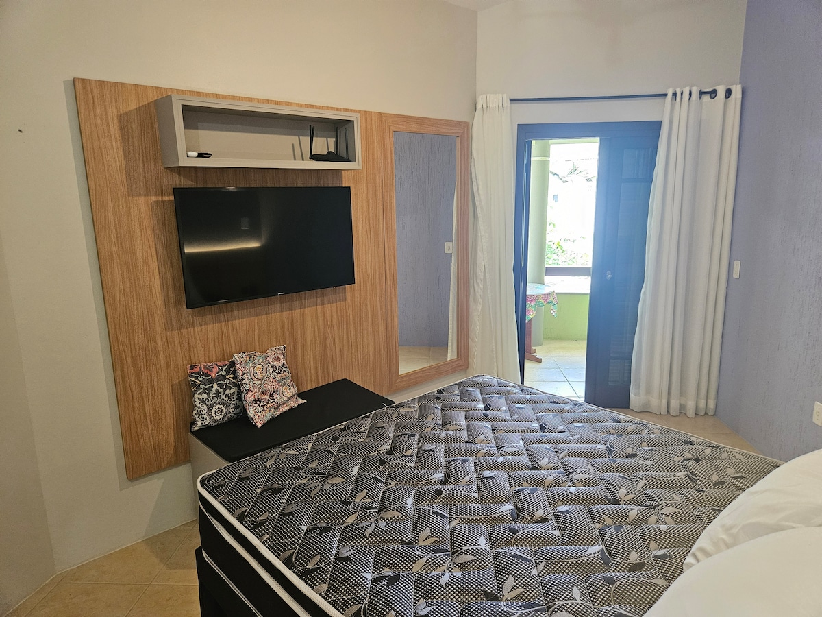 A cozy integrated living space is showcased, featuring a queen bed with a patterned mattress and decorative pillows. A wall-mounted television is visible above a black bench. Natural light enters through the balcony door leading to an outdoor area.