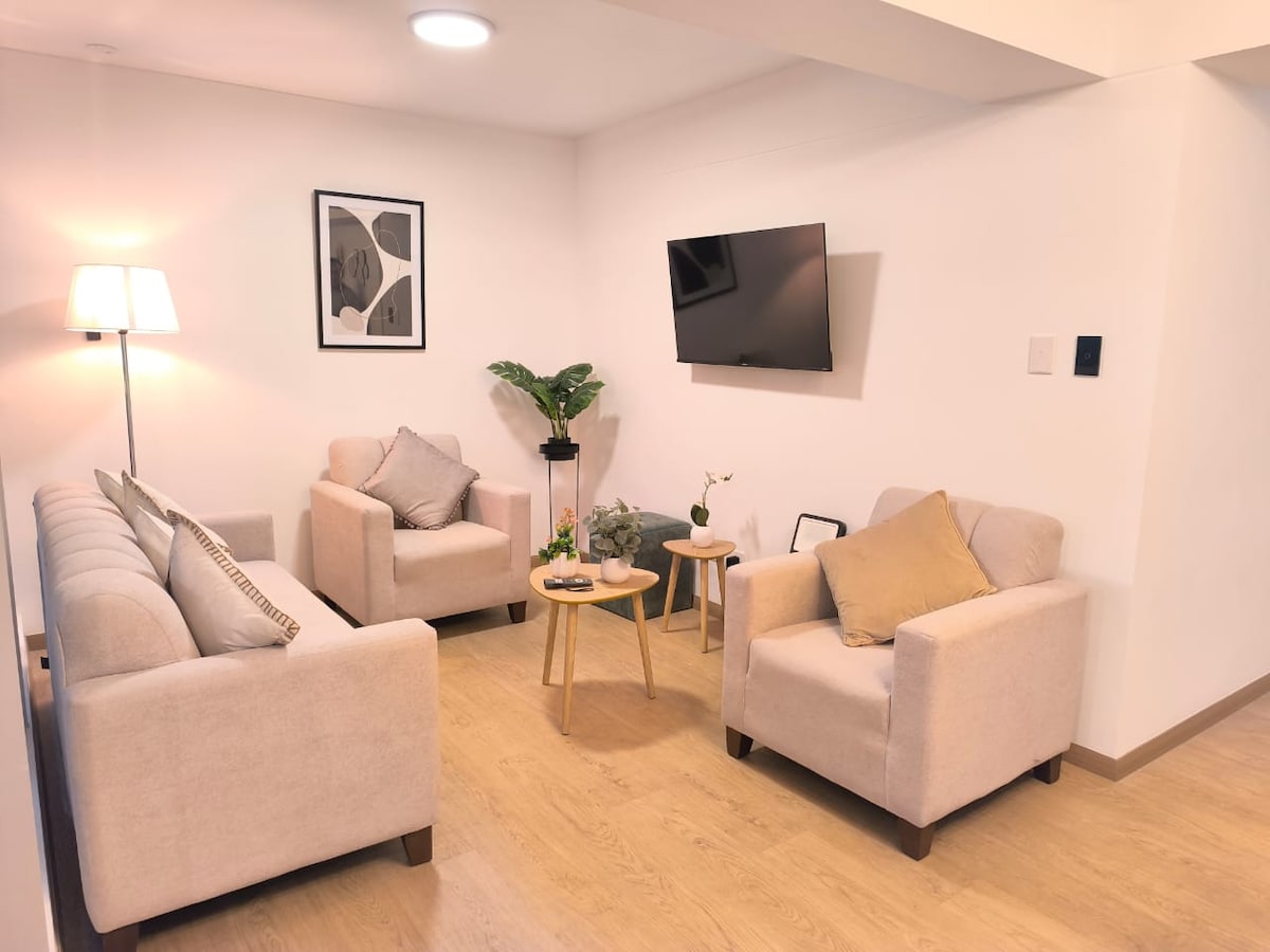 A contemporary living area is furnished with a comfortable beige sofa and two armchairs. A small wooden coffee table is centered among them, displaying decorative items. A wall-mounted television is positioned across from the seating. Soft lighting creates a cozy ambiance, complemented by a potted plant.