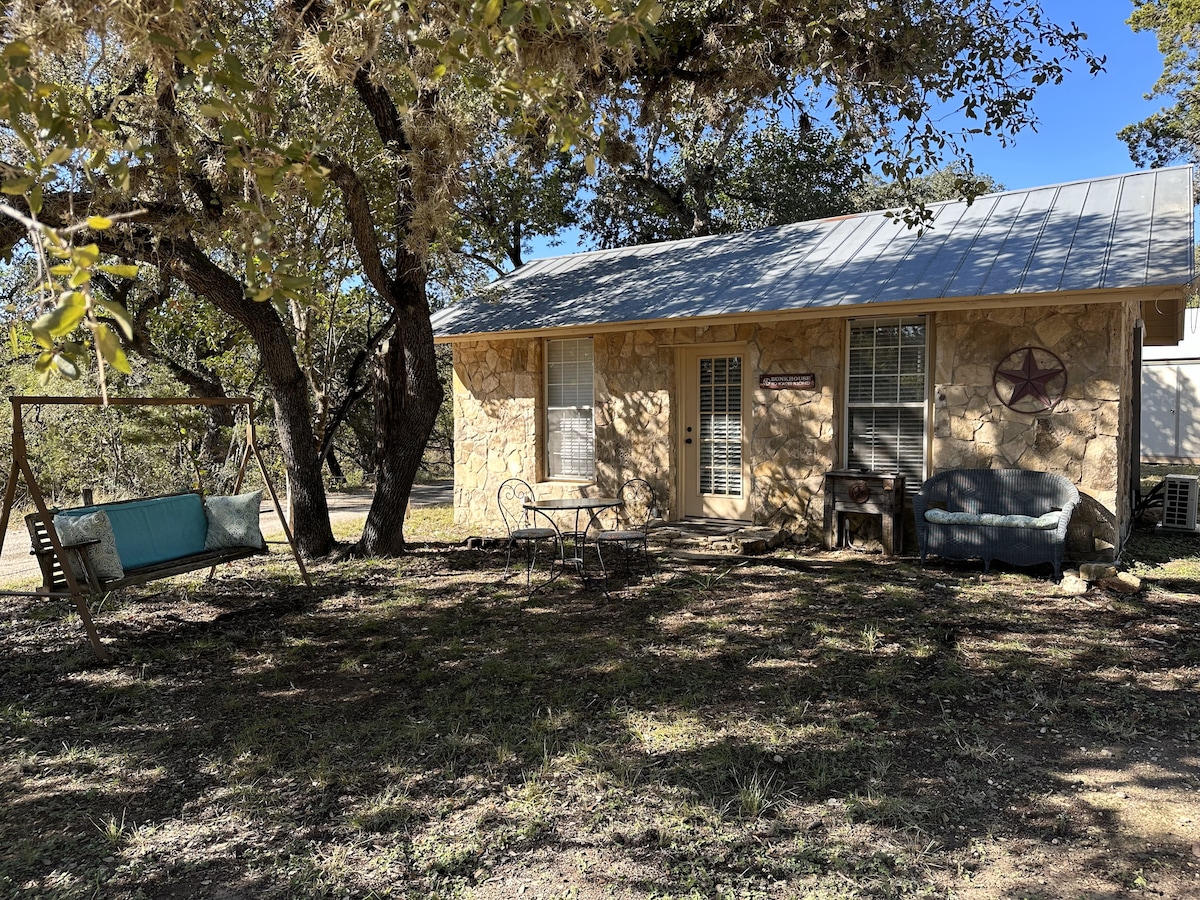 The exterior of The Bunkhouse is framed by a large live oak tree, providing shade. A swing hangs from the tree alongside a small table and chairs set, with a rustic bench nearby. Stone walls and a simple metal roof enhance the inviting outdoor space.