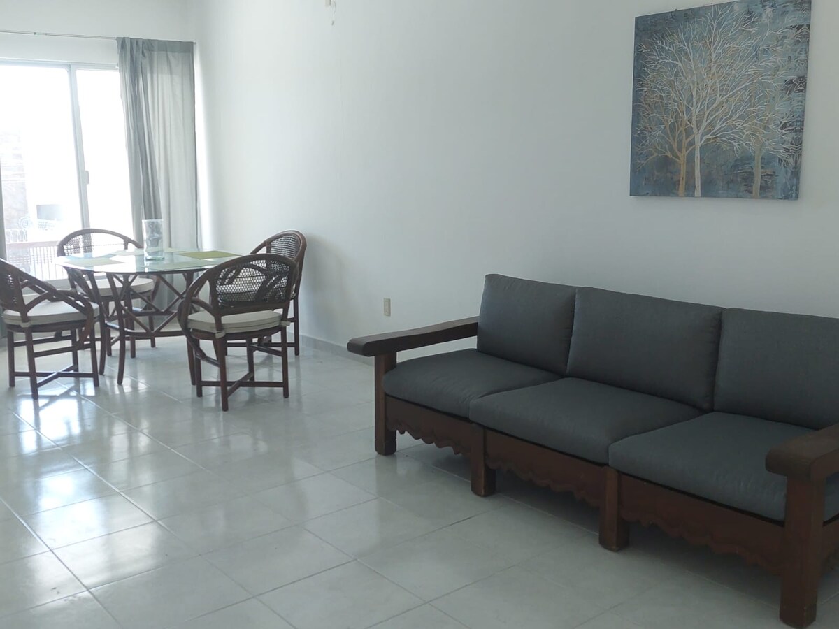 A bright living area features a comfortable gray sofa without armrests, accompanied by a glass dining table surrounded by four rattan chairs. Light streams in through large windows with sheer curtains, illuminating the white tiled floor and a wall art piece depicting a tree.