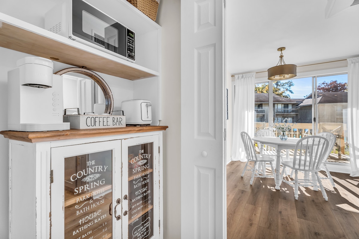 A compact kitchen area is shown, featuring white cabinetry and a wooden countertop. A coffee station is neatly arranged with a coffee maker and toaster. In the background, a bright dining space is visible, showcasing a round table surrounded by white chairs, illuminated by ample natural light.