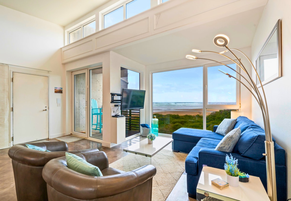 The living room features comfortable seating with a blue sectional sofa and two brown armchairs. A sleek coffee table sits at the center, while large windows showcase ocean views and allow natural light to fill the space. A TV is mounted on the wall, enhancing the relaxation atmosphere.