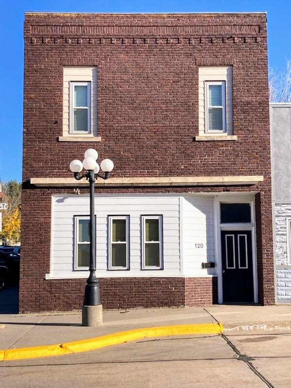 A brick building with a white accent facade is featured, showcasing large windows framed in white. A decorative lamppost stands at the forefront, illuminating the entrance. The building is set on a main street, highlighting its urban environment.