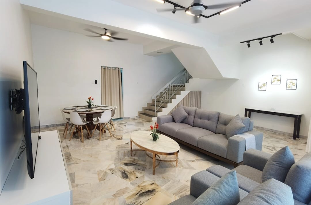 A spacious living area is presented, featuring a large sectional sofa and a round coffee table. A flat-screen television is mounted on the wall. Natural light fills the room, highlighting the marble flooring. A dining area with six chairs is positioned nearby, complemented by a staircase in the background.