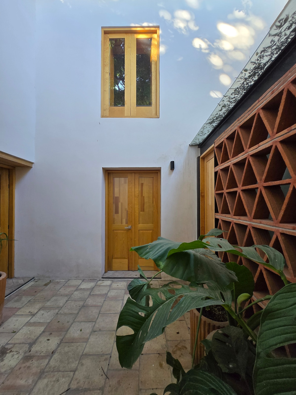 A serene courtyard features textured clay walls and warm wooden doors. Sunlight filters through a tall window, creating delicate shadows on the tiled floor. Lush greenery adds a natural element, enhancing the calm ambiance of the space.
