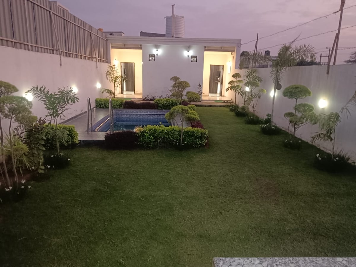 A well-maintained garden surrounds a tranquil swimming pool, enhanced by neatly trimmed hedges and ornamental plants. Soft lighting highlights the landscape, while two entry doors can be seen leading to the villa. A clean, manicured lawn adds to the serene atmosphere.