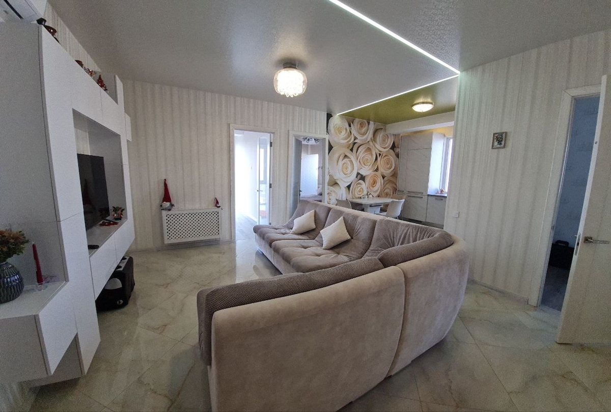 A spacious living area features a light-colored sectional sofa with decorative cushions. A wall-mounted television is placed on a modern white cabinet. Subtle lighting fixtures are present, while a floral-patterned accent wall adds a touch of charm to the room.