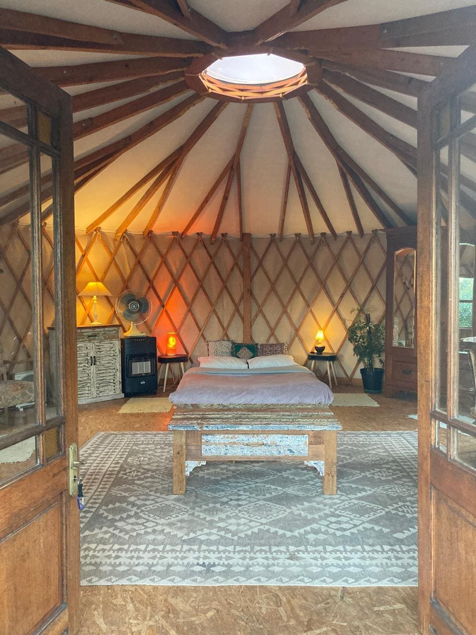 The interior of the yurt presents a cozy sleeping area featuring a spacious bed at the center, framed by wooden walls with intricate lattice designs. A wooden table sits in front of the bed, complemented by decorative lighting and a fan. Natural light filters through a circular skylight.