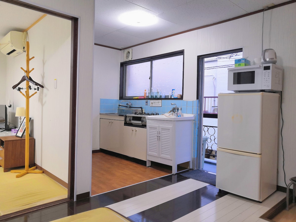 A compact kitchen area is visible, featuring a sink, stove, and a microwave atop a white cabinet. A refrigerator stands nearby, and a window allows natural light to brighten the space. The room connects to a cozy sleeping area, adorned with simple furnishings.
