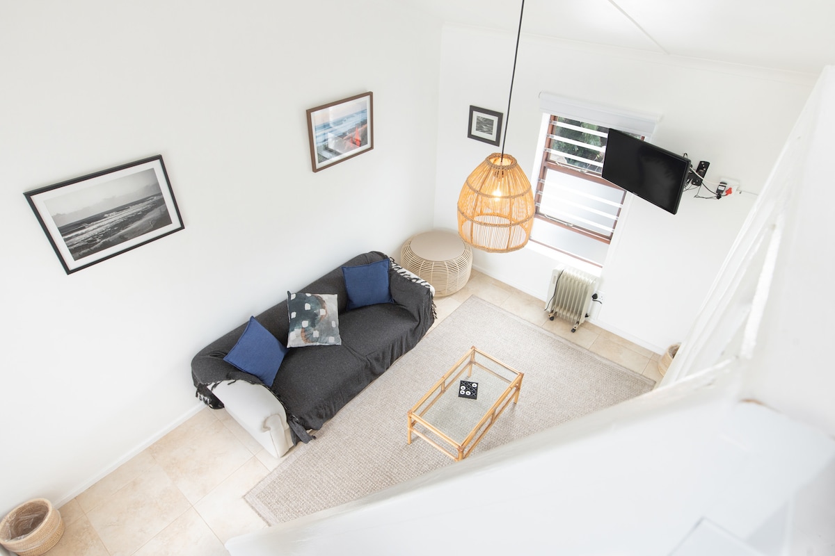 A bright lounge area is showcased with a cozy sofa adorned with decorative pillows. A rattan coffee table is positioned on a neutral rug, complemented by framed ocean photography on the walls. A television is mounted near a window, allowing natural light to illuminate the space.