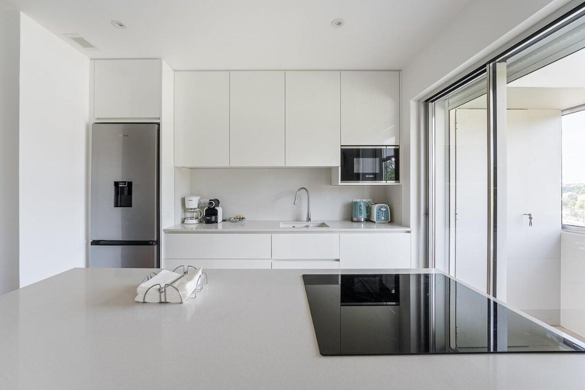 A modern kitchen is visible, featuring sleek, white cabinetry and a spacious countertop. High-end appliances, including a refrigerator and a microwave, are present. Natural light fills the area through large windows that open to a balcony, creating a bright and open cooking space.