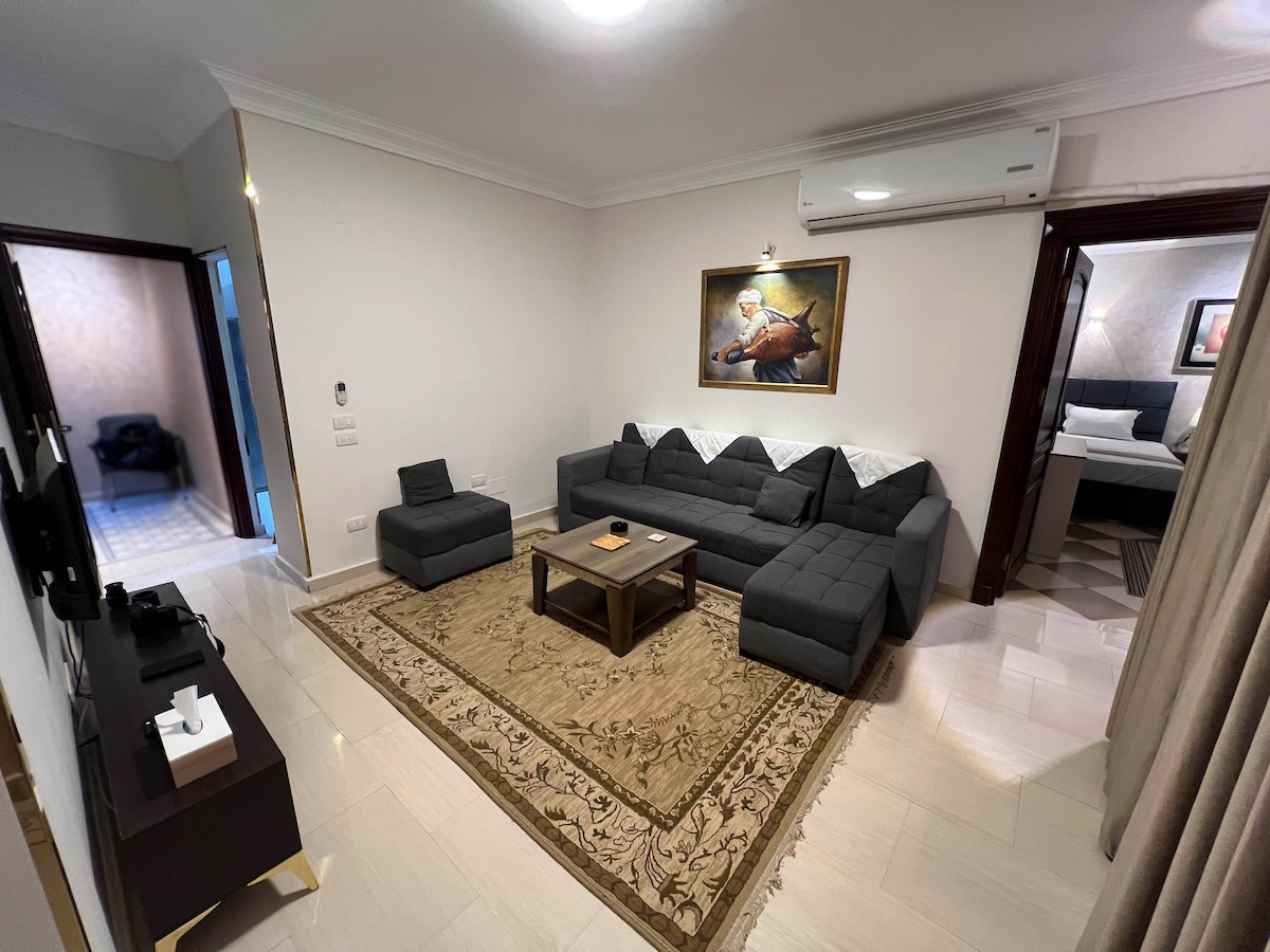 A spacious living area is displayed, furnished with a gray sectional sofa and a coffee table. Artwork adorns the wall, and an air conditioning unit is mounted above. Light-colored flooring complements the decorative area rug, adding to the room's inviting ambiance.