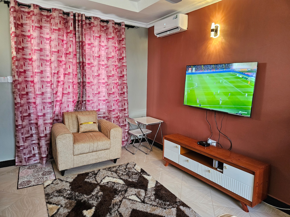 A cozy living area is highlighted by a comfortable armchair and a modern TV mounted on the wall. An accent rug adorns the tiled floor, and a small table is positioned nearby. Warm-colored curtains frame the window, adding a touch of softness to the space.