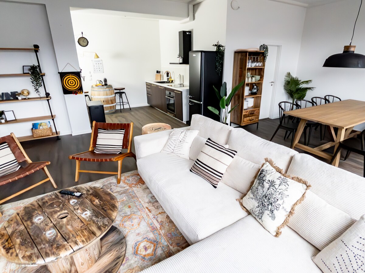 A spacious living area is highlighted by a large, cream-colored sectional couch adorned with decorative pillows. A rustic coffee table sits in the center, while two wooden lounge chairs provide additional seating. The background features an open kitchen and a dining area with a wooden table.