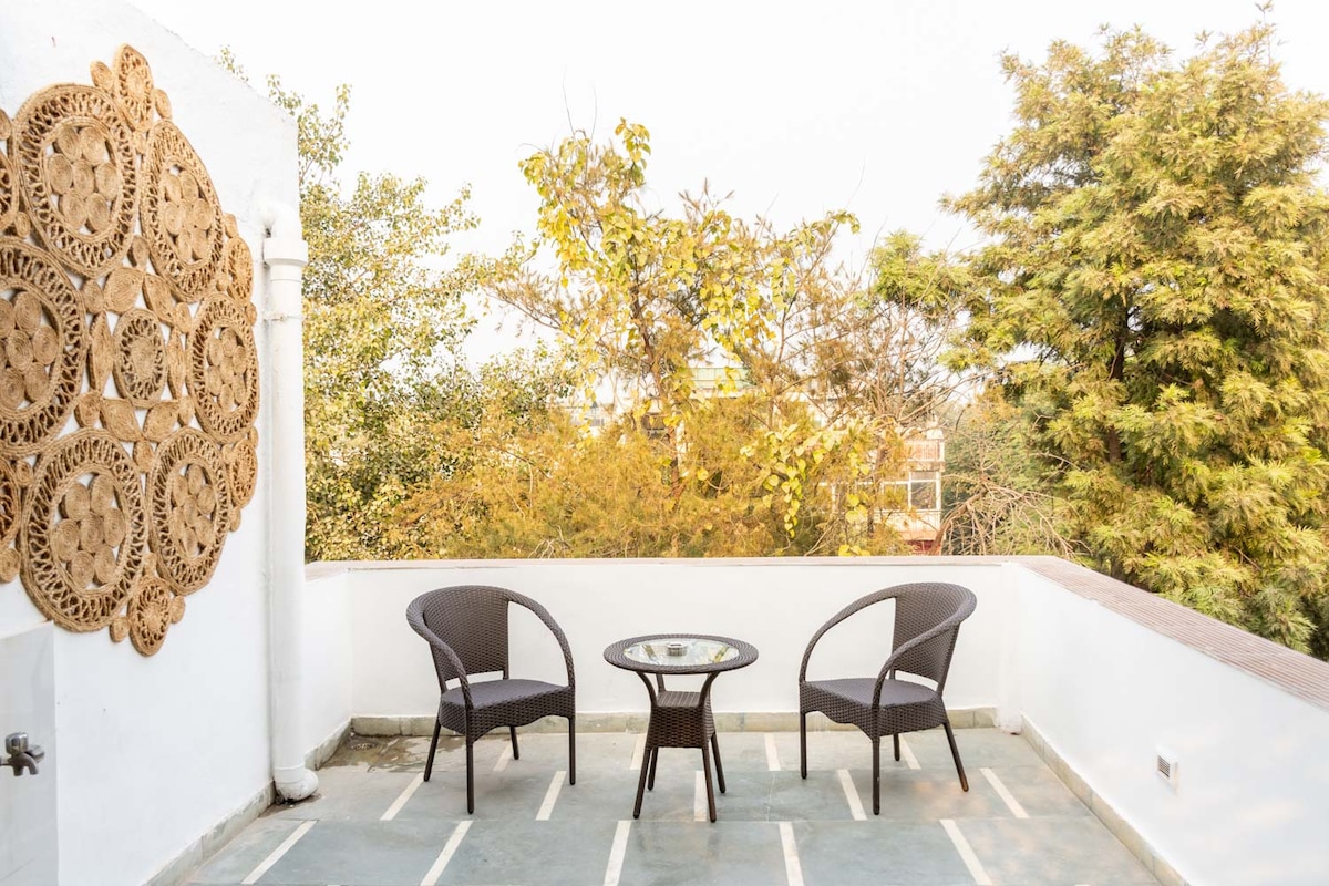 A serene balcony space features two woven chairs paired with a glass table, surrounded by lush greenery. Decorative wall accents enhance the atmosphere, while the natural light creates a bright and inviting environment for relaxation.