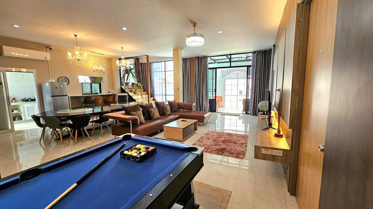 A spacious living area features large windows that provide ample natural light. A billiards table is positioned in the foreground, while a comfortable seating arrangement and a dining table are visible in the background. The open layout enhances the sense of flow between spaces.