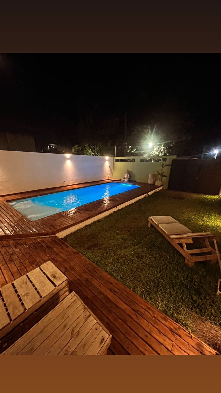 An inviting outdoor space features a swimming pool illuminated with soft blue lighting, surrounded by wooden decking. A well-maintained lawn extends beside the pool, complemented by minimalist lounge chairs positioned for relaxation.