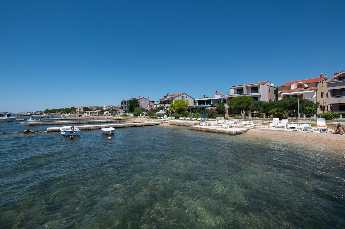 A serene coastal view features a sandy and pebble beach lined with white lounge chairs. Nearby, boats can be seen floating on clear turquoise water, with residential buildings positioned along the shore, surrounded by greenery under a clear blue sky.