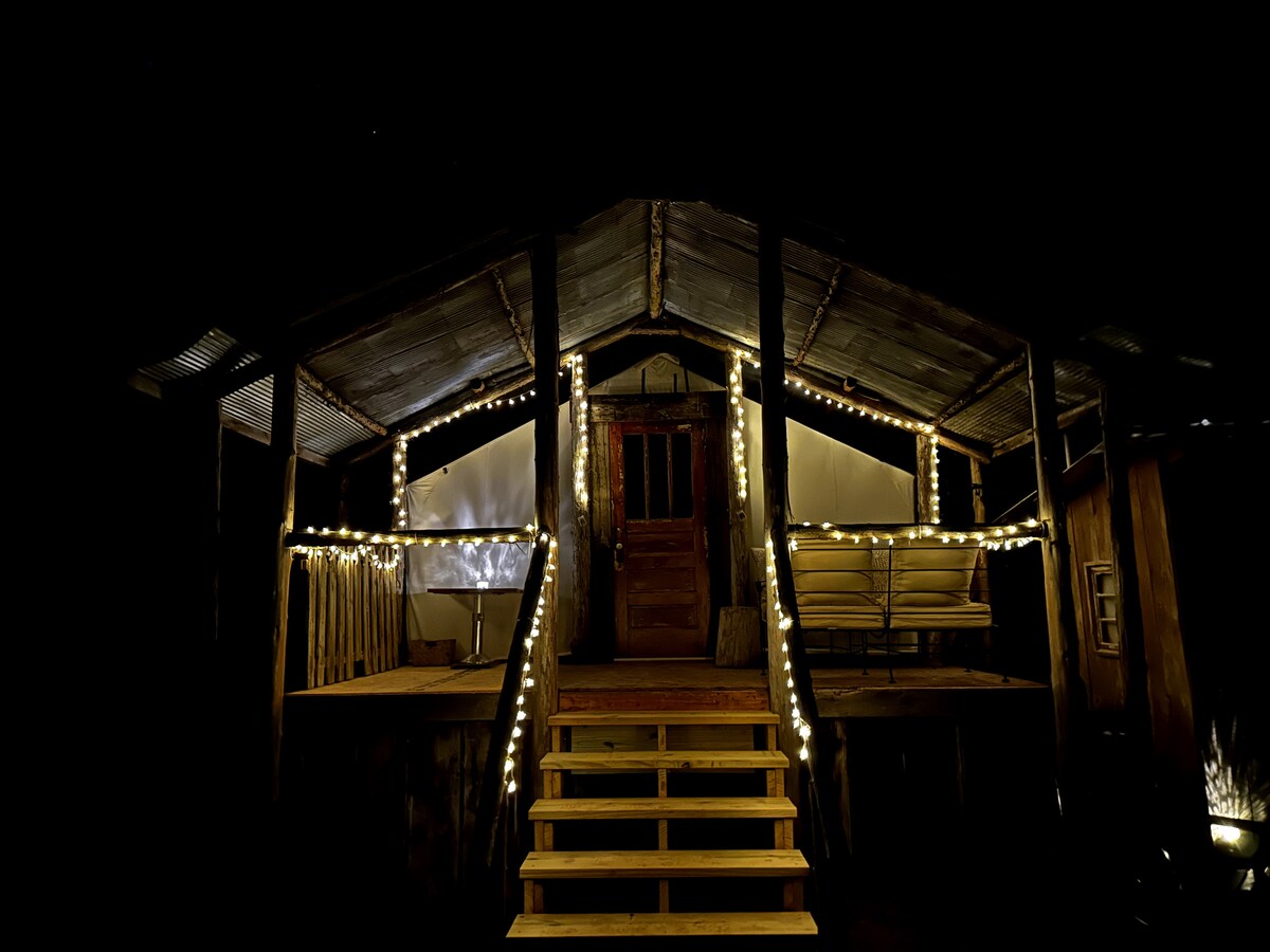 The tentalow's exterior is illuminated by string lights, highlighting the rustic charm of its wooden structure. Stairs lead up to the entrance, flanked by a cozy seating area on the porch. The night sky serves as a backdrop, enhancing the inviting ambiance.