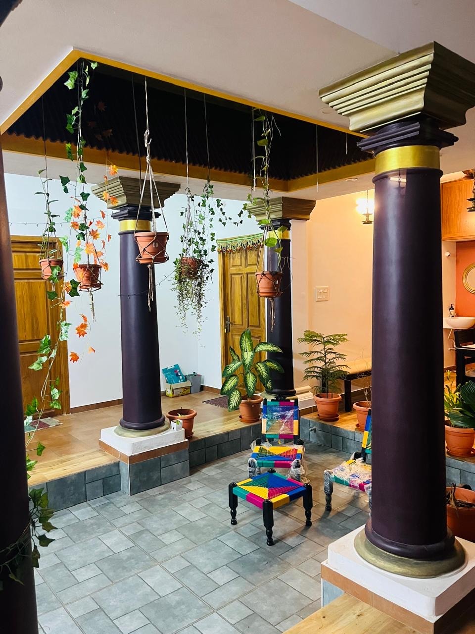 A vibrant entryway is defined by sturdy columns adorned with decorative finishes. Hanging plants and flower pots create a lively atmosphere. Colorful chairs are positioned around a tiled floor, with potted greenery placed throughout the space, contributing to a refreshing indoor garden feel.