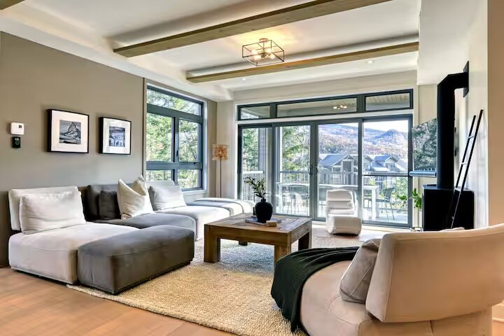 The living area features a spacious sectional sofa in muted tones, complemented by a wooden coffee table. Large glass doors allow natural light to fill the space, leading to a scenic balcony view. Artwork adorns the walls, adding subtle character to the neutral decor.