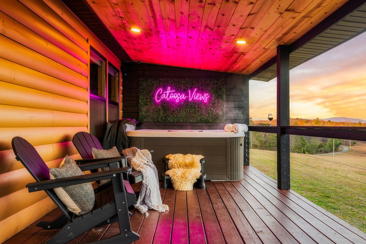 A wooden deck features a 4-person hot tub surrounded by lush greenery and neon signage reading 'Catoosa Views.' Two Adirondack chairs are positioned nearby, accompanied by a cozy throw blanket and decorative pillows, with stunning sunset colors visible in the background.