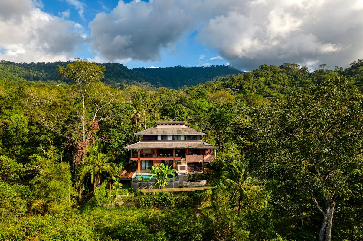 A three-level home is nestled among lush greenery, surrounded by towering trees and mountains in the background. The building features a blend of natural elements and modern design, with expansive outdoor areas that enhance the connection to nature.