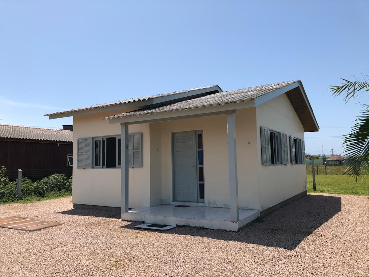 A single-story structure is presented with a light exterior and a gabled roof. Shuttered windows enhance the facade, while a covered entrance offers shelter. The gravel path leads to the front door, surrounded by open green space and a few distant buildings.