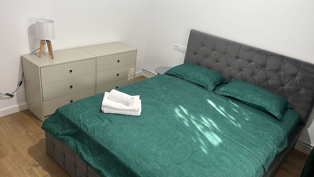 A spacious bed is dressed in rich teal bedding, complemented by neatly folded white towels. A minimalist dresser with multiple drawers and a sleek nightstand are positioned nearby. Soft light filters through the room, highlighting the clean lines of the modern decor.