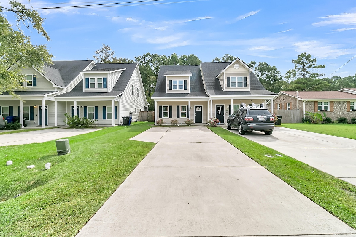 A well-maintained duplex is presented, featuring a spacious driveway suitable for parking. The front yard is landscaped with green grass and a few shrubs. The two-story home displays a welcoming facade with a covered entry and multiple windows.