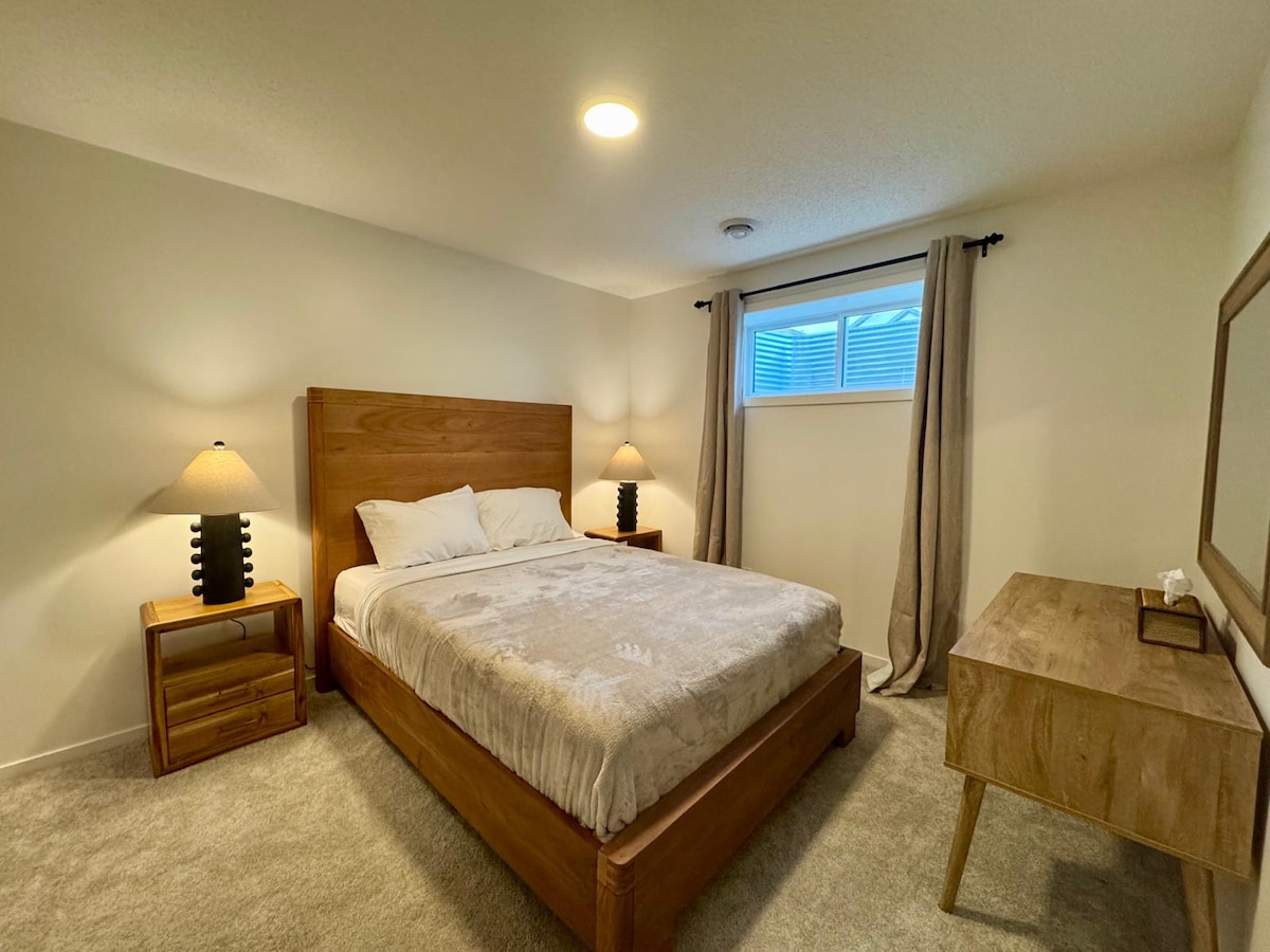 A well-furnished bedroom features a spacious bed with soft linens, flanked by two bedside lamps. Natural light floods the room through a window adorned with light curtains. A wooden dresser offers additional storage, enhancing the simple and functional design.