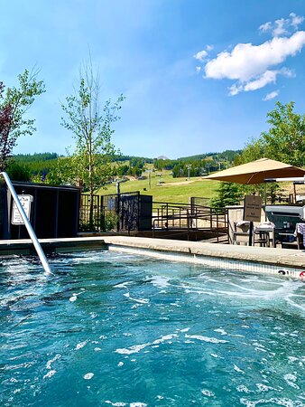 A clear blue sky is visible above a sparkling hot tub located near a scenic area. Surrounding greenery and the gentle slopes in the background create a serene outdoor space. A shaded seating area is present nearby, offering relaxation options.