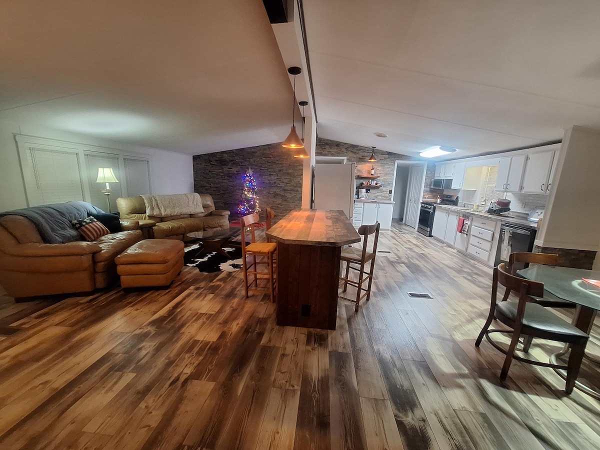 A spacious living area features warm wooden flooring and is divided into a seating section with a large sectional sofa and a dining area with a wooden bar counter and several chairs. Soft lighting enhances the cozy ambiance, complemented by stone accent walls.