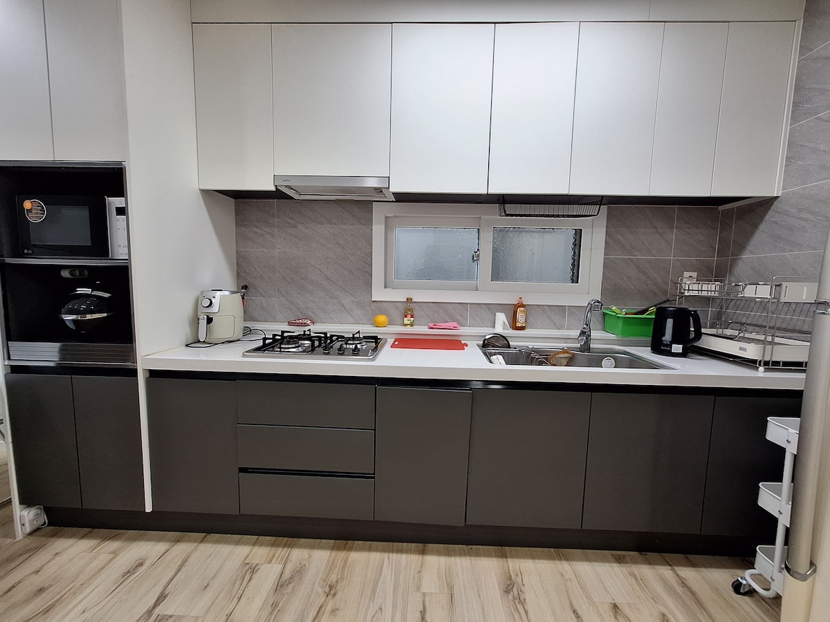 A modern kitchen space features sleek cabinetry with a combination of light and dark tones. Equipped with a gas stove, microwave, and a sink, the area is organized with various cooking essentials, complemented by a window allowing natural light. Appliances are neatly arranged for convenience.