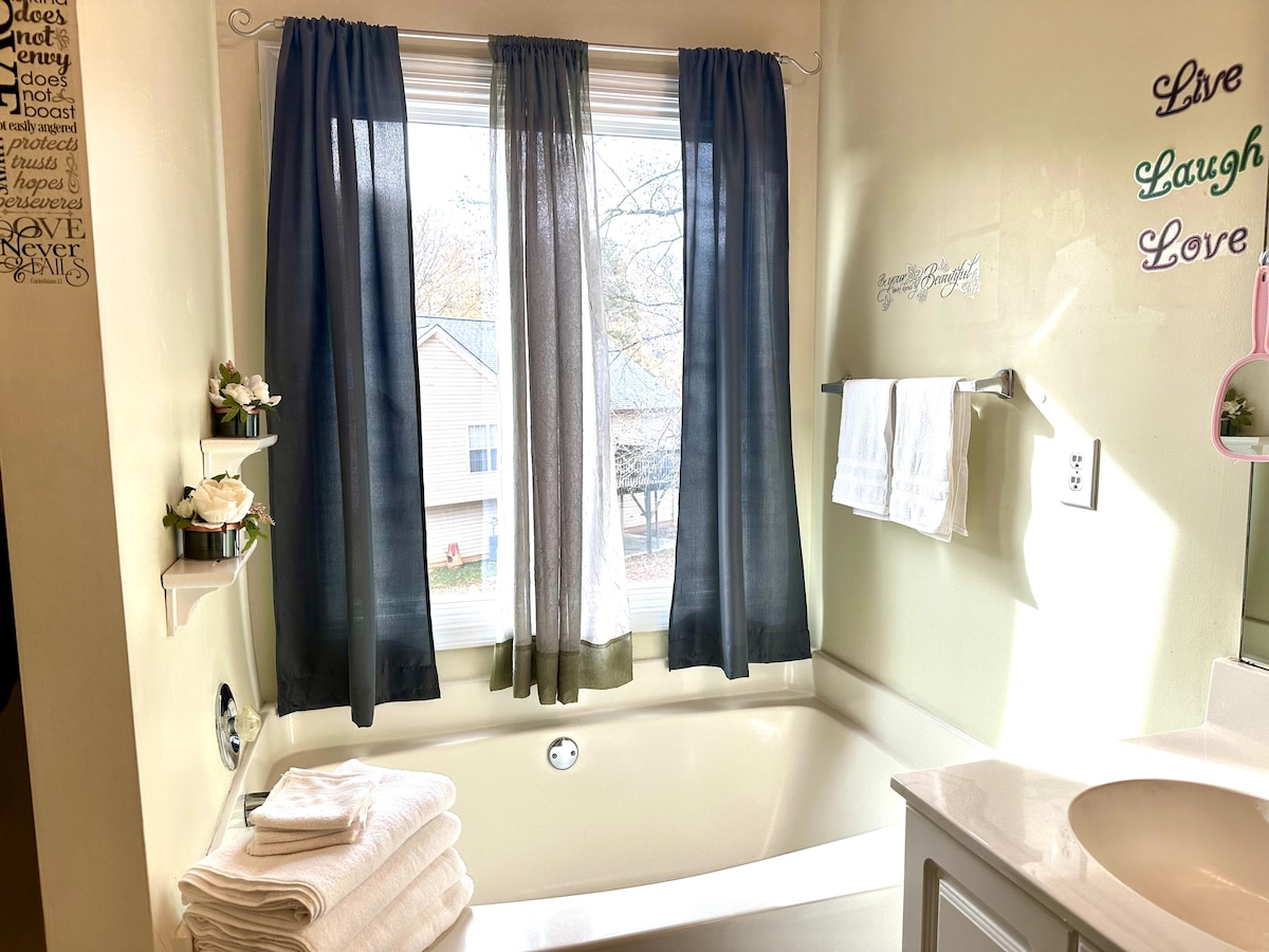 A well-lit bathroom features a large soaking tub beneath a window adorned with sheer and dark curtains. Neatly stacked towels and decorative shelves with greenery are visible, contributing to the serene atmosphere. The sink area is complemented by reflective surfaces and soft natural light.