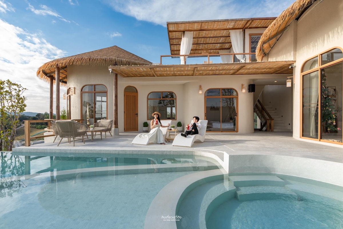 A spacious outdoor deck is shown featuring relaxing lounge chairs beside a private pool. The villa’s large windows and bamboo roofing create a harmonious blend with the surrounding landscape. Two guests are seated, enjoying the serene ambiance and picturesque views of the hills.