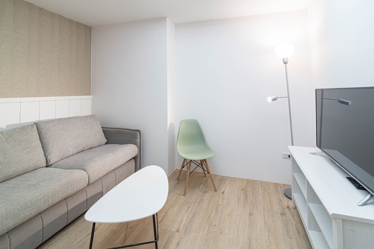 A modern living area is depicted, featuring a light gray sofa paired with a minimalistic white coffee table. An accent chair in pastel green adds a touch of color. A lamp provides soft lighting, while a flat-screen television rests on a simple white console.