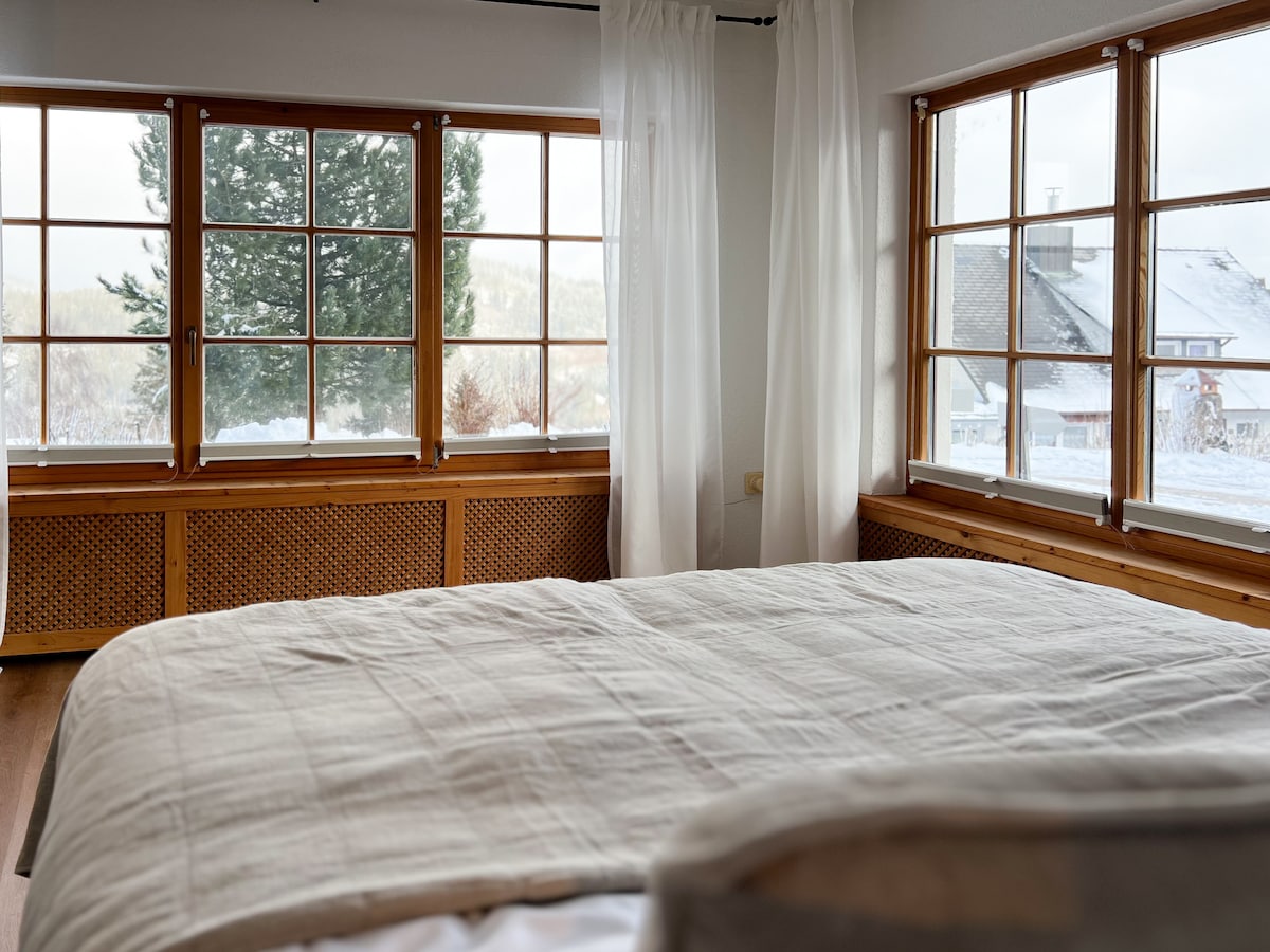 A well-lit bedroom is featured, showcasing a spacious bed covered with a light-colored quilt. Large windows framed in wood allow natural light to fill the room, with views of snow-covered outdoor scenery and a green tree visible outside.